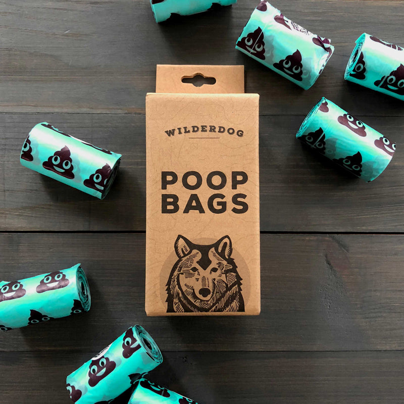 A box labeled "Dog Poop Bags" with a wolf illustration is surrounded by extra-thick rolls featuring cartoon poop emojis, all arranged on a dark wooden surface.